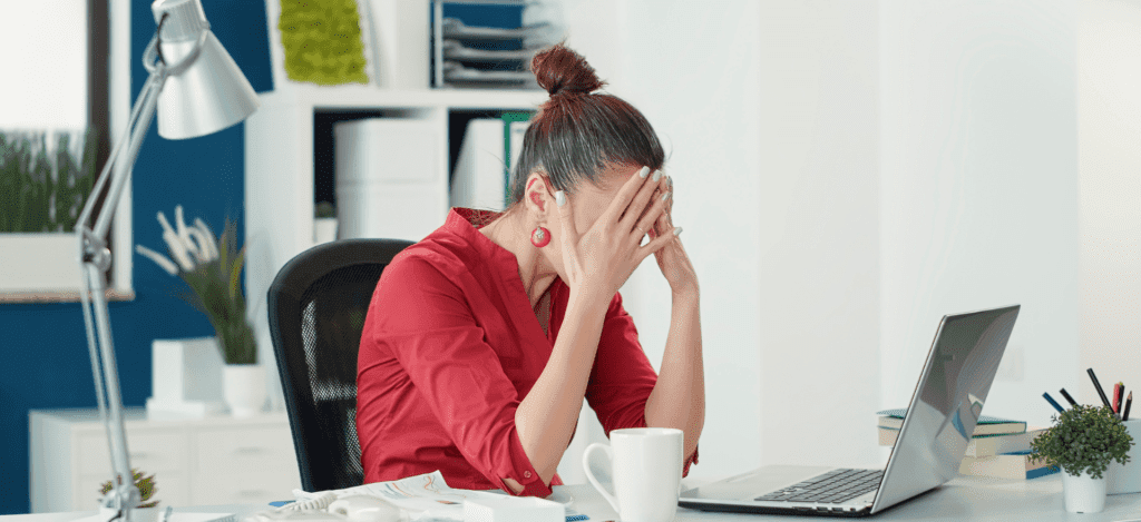 A woman in a red shirt sitting at a desk with her hands covering her face in frustration, symbolizing the stress caused by credentialing mistakes and administrative burdens.